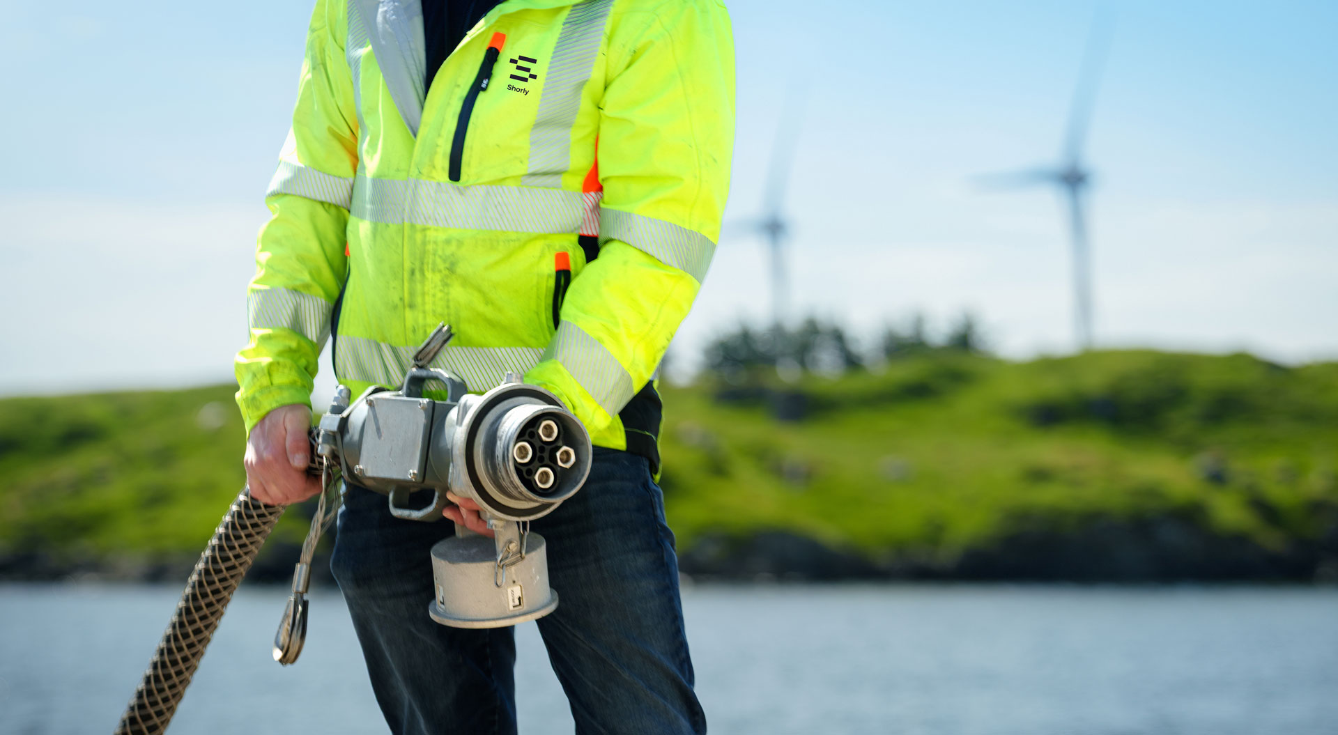 A Shorly technician in a high-visibility jacket carrying a shore power cable with wind turbines in the background